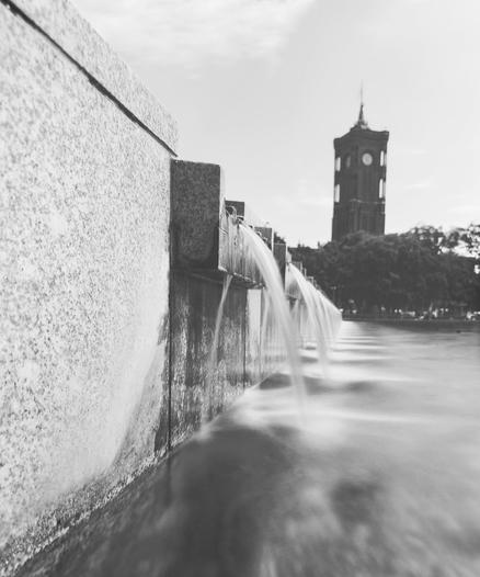 A long exposure of water flowing over a cascade. The water has a semi-silkiness to it.  In the background are some trees and beyond that the tower of the Rotes Rathaus (Red Town Hall).  This is a black and white image and the back ground is a stark black.